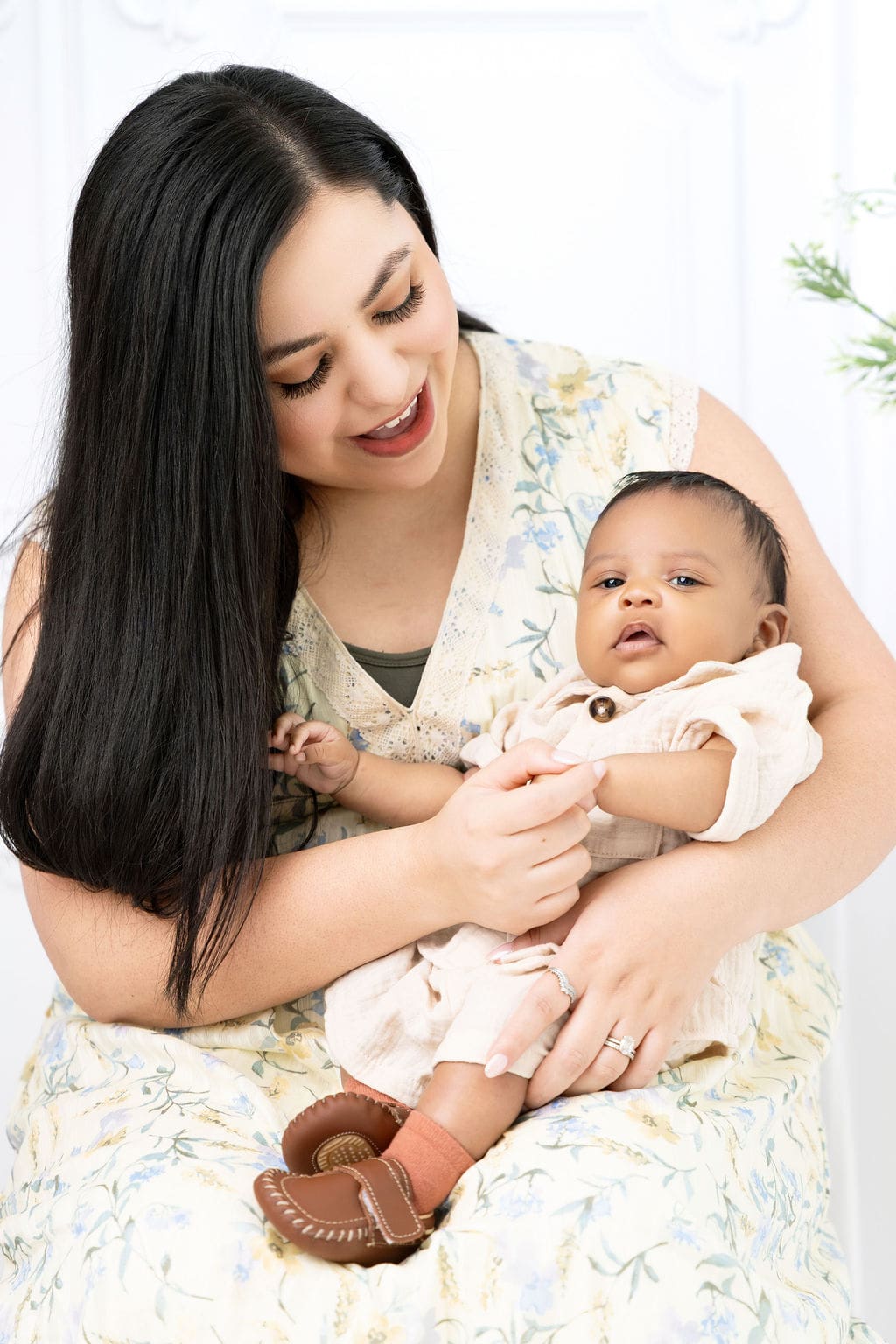 A happy baby sits in mom's lap wearing a cream onesie and leather shoes as mom plays with him