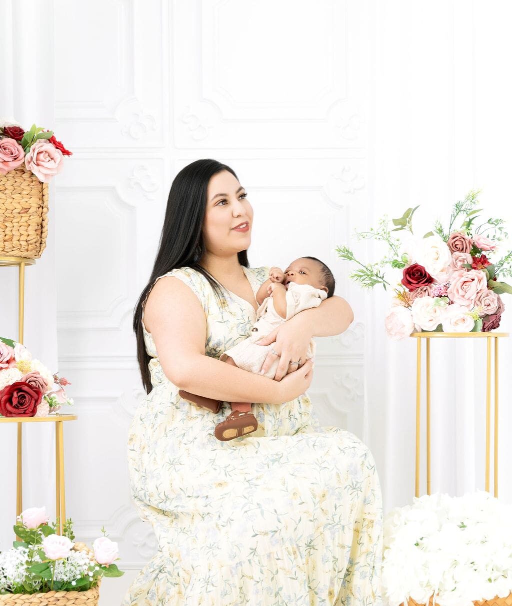 A happy new mom in a yellow floral print dress sits in a studio surrounded by roses holding her infant in her arms after finding great baby stores in austin