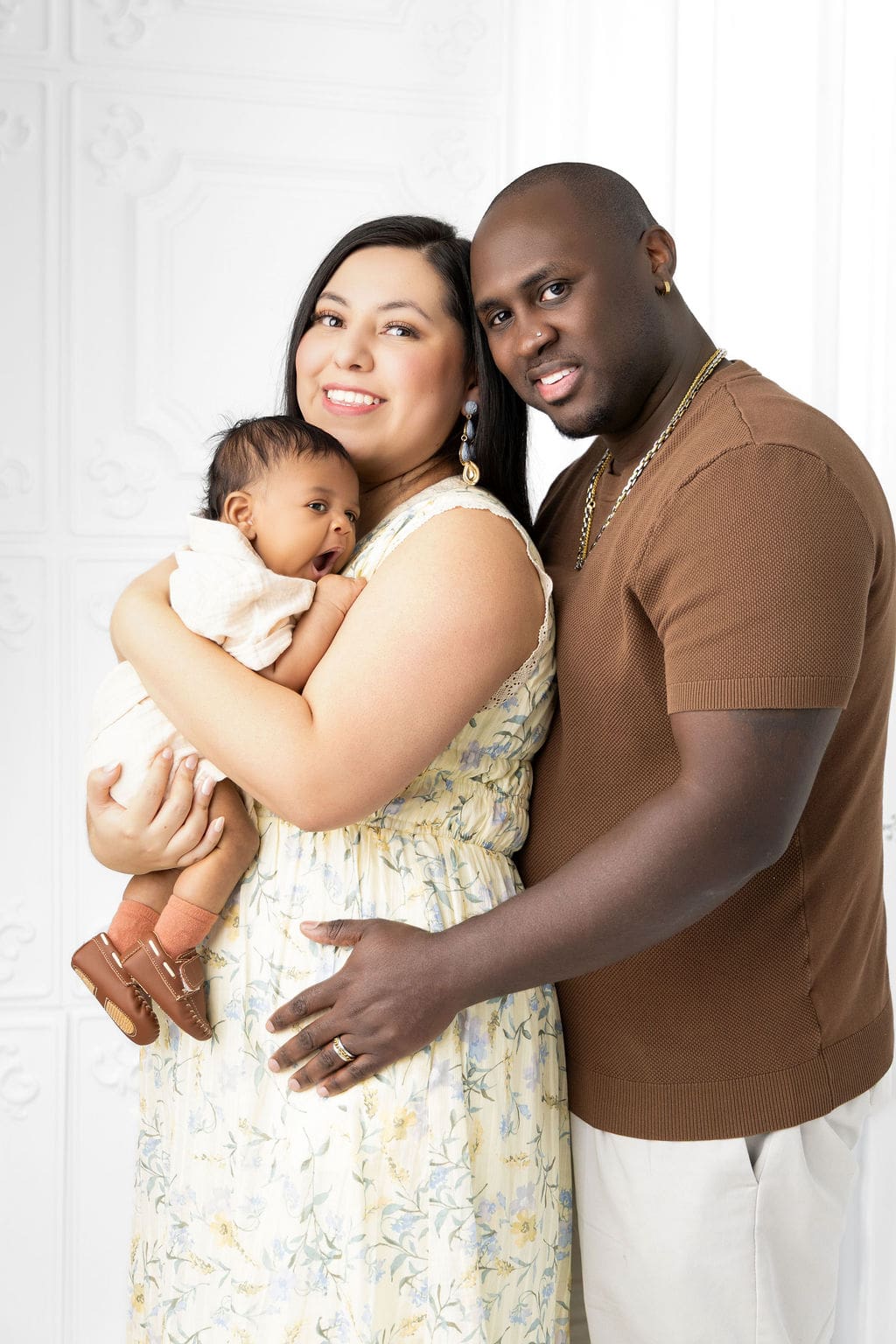 Happy new parents stand in a studio holding their yawning baby against mom's chest in a floral print dress and brown shirt after visiting baby stores in austin