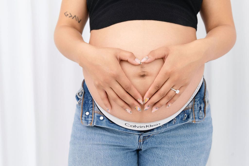 A woman holds her hands in the shape of a heart on her pregnant bump while wearing open jeans and a black blouse