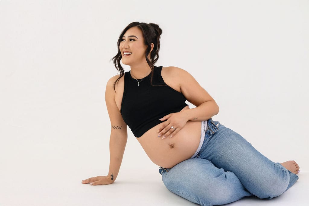 A smiling mom to be sits on the floor of a white studio with a hand on her bump and smiling off to the distance after meeting doulas in austin in a black top and jeans
