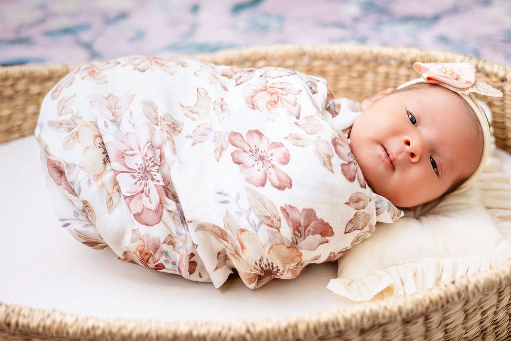 A newborn baby girl in a flower print swaddle lays in a woven basket with eyes open