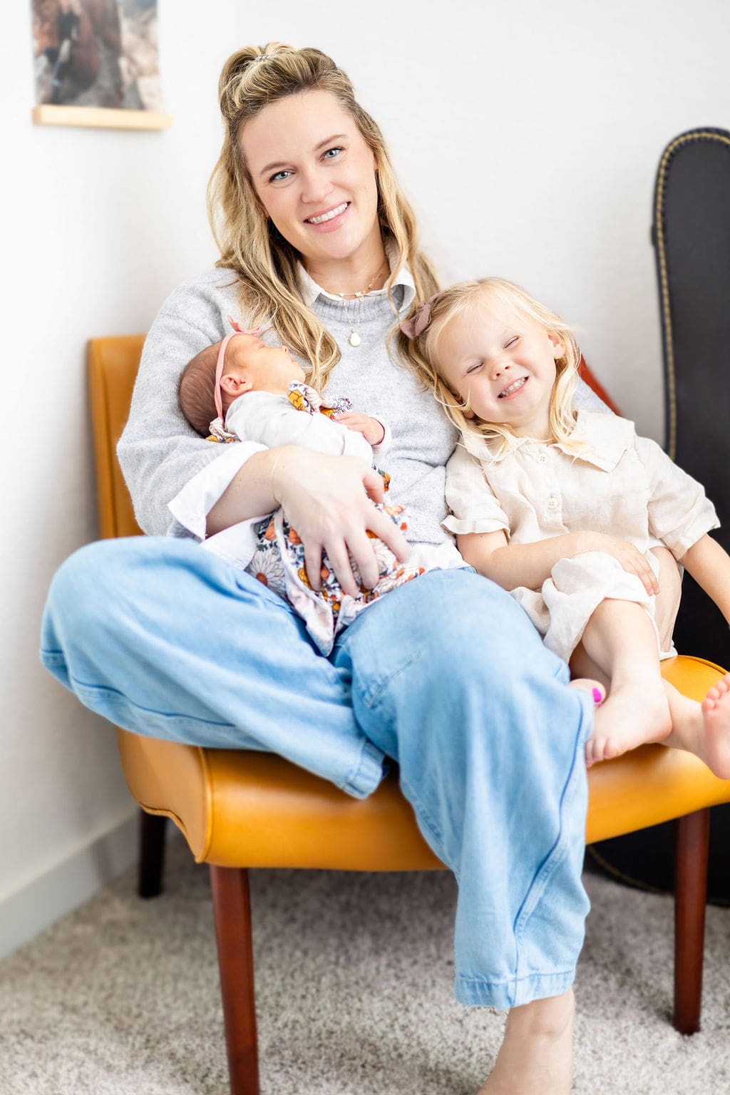 A happy mom sits in a leather chair holding her sleeping newborn and smiling toddler in her lap after meeting lactation consultants in austin