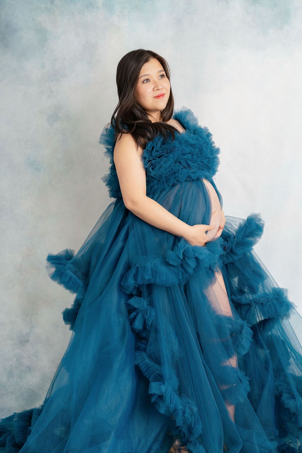 A happy mother to be stands holding her bump while in a blue tulle maternity gown in a studio