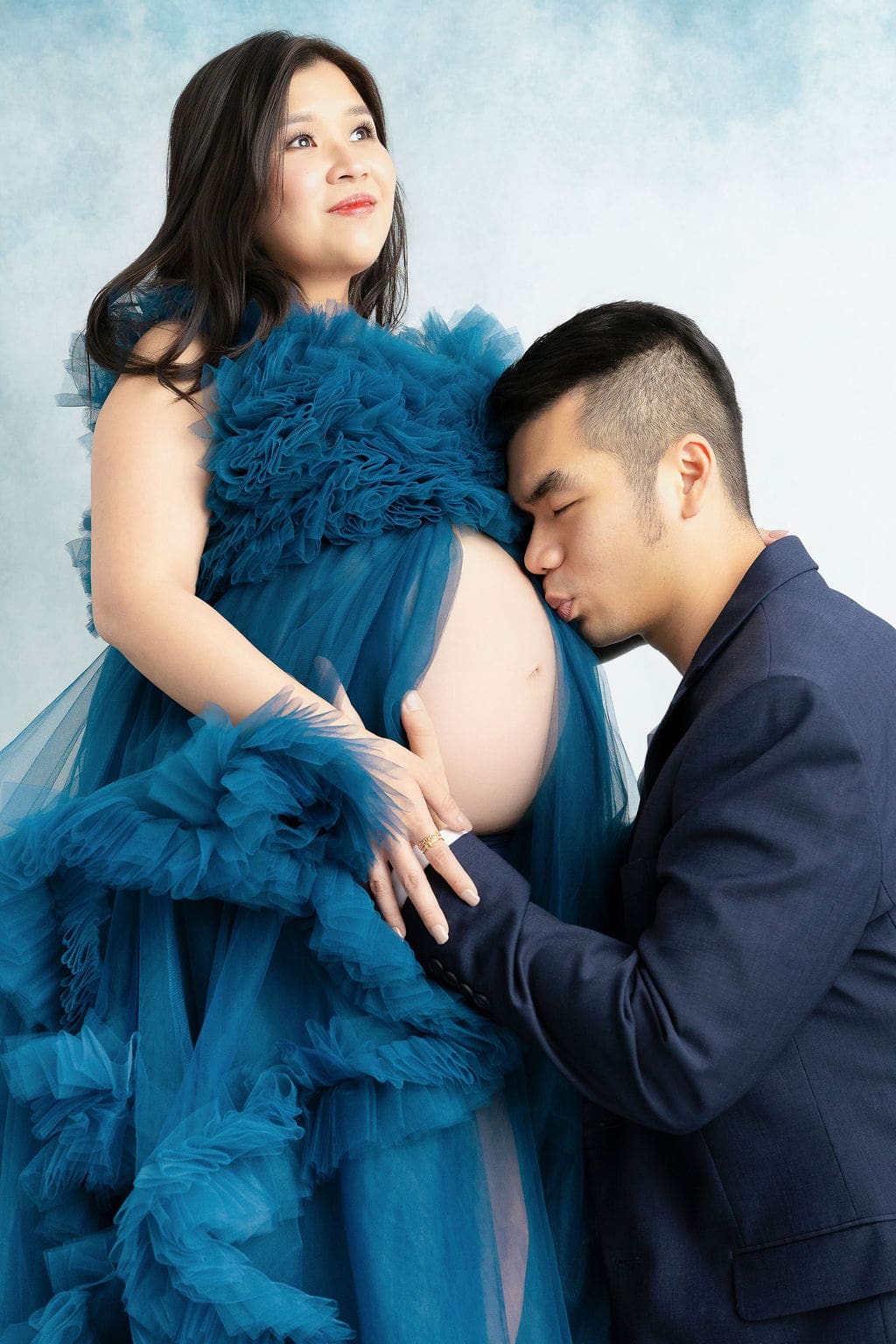 A pregnant woman stands in a studio in a blue maternity gown as her husband in a blue suit jacket kisses the bump after meeting midwives in austin