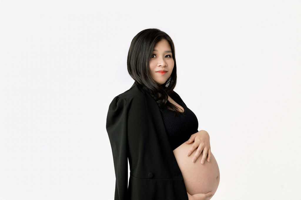 A mom to be stands holding her bump while looking over her shoulder in a white studio with a black sweater on her shoulders