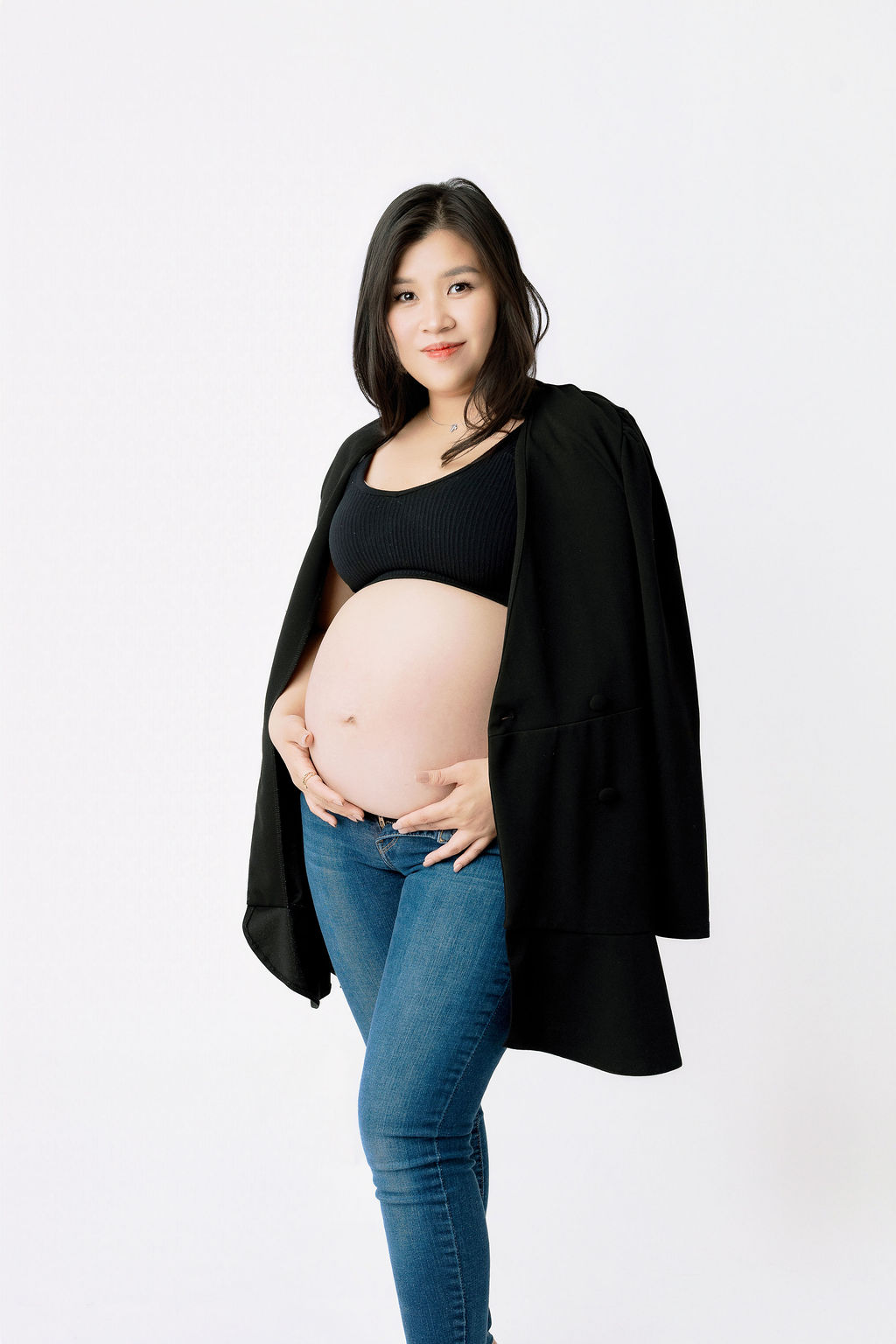 A mother to be in an open black sweater and jeans stands smiling with hands under her bump in a studio after a 3d ultrasound in austin