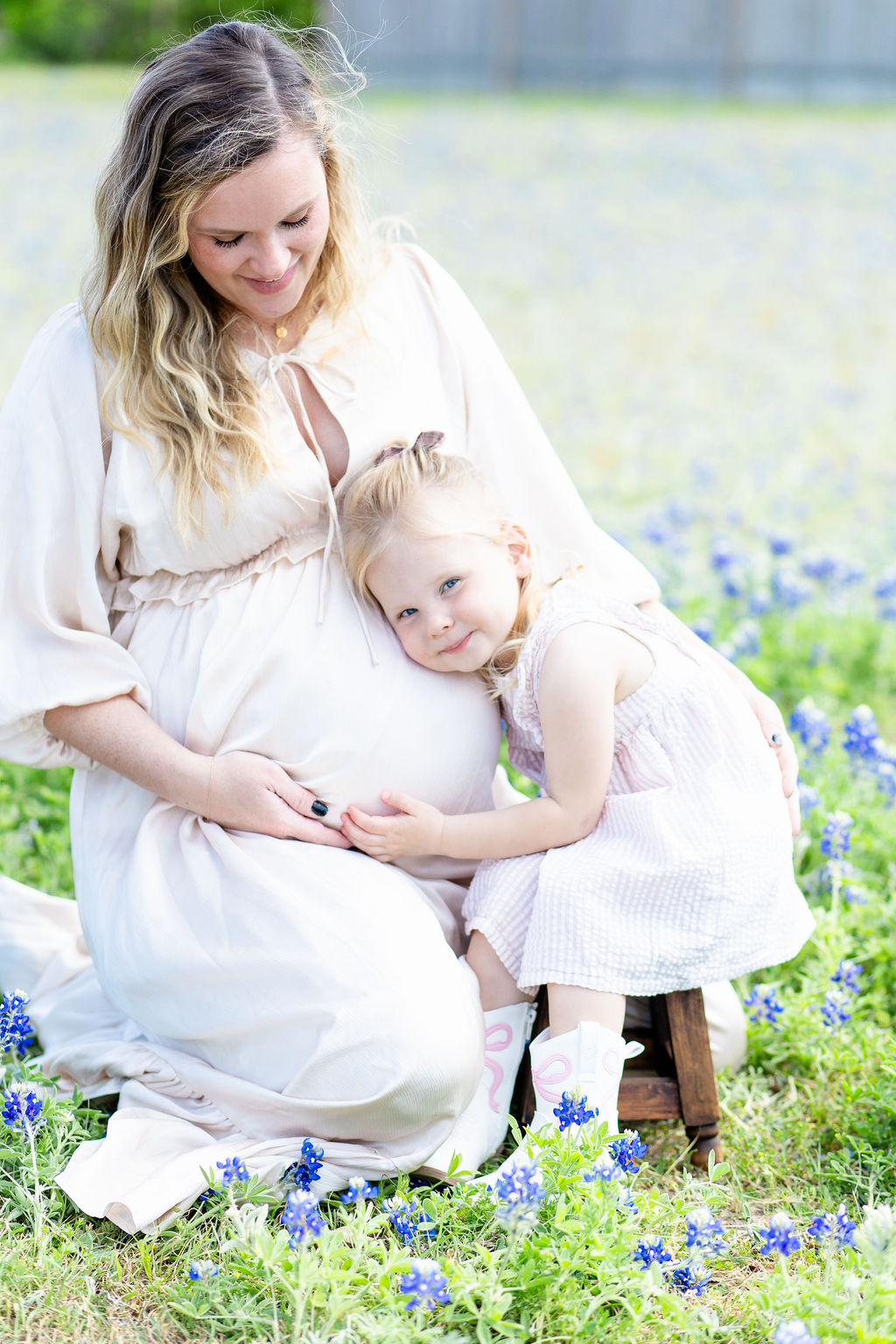 A happy pregnant mother in cream is hugged by her toddler daughter in a field of blue flowers