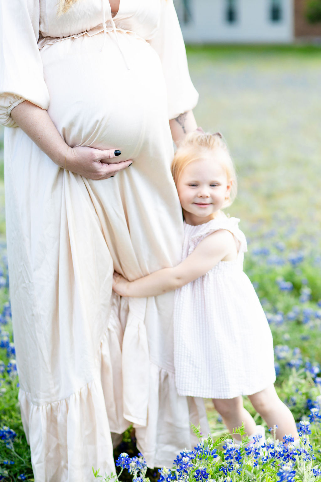 A toddler girl in white hugs her pregnant mom's leg in a field of blue wildflowers while mom holds the bump after visiting birth centers in austin