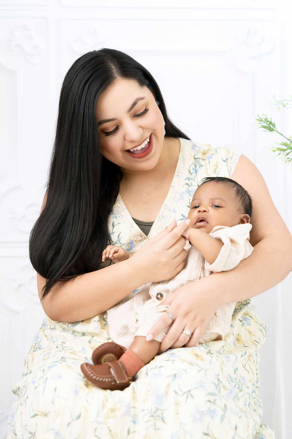 A happy new mom in a yellow dress plays with her tired infant in her lap while sitting in a studio