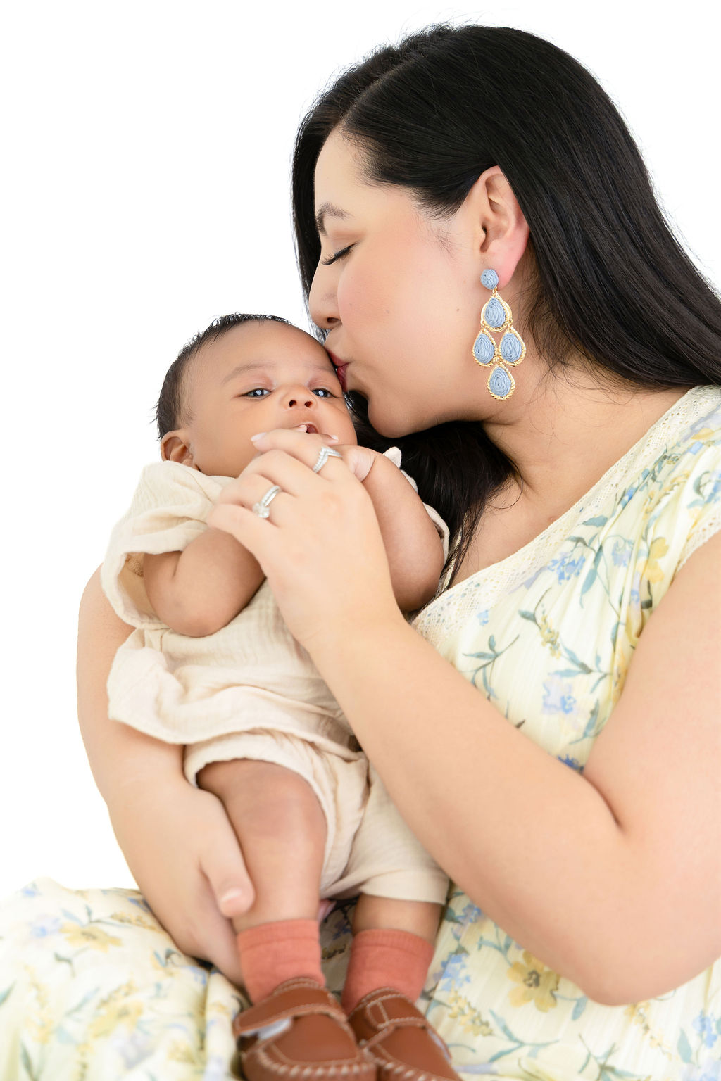 A happy new mom in a yellow floral dress sits while kissing her infant in her arms after meeting with nannies in austin