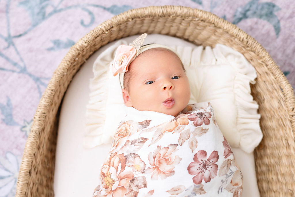 A newborn baby makes a silly face while awake in a woven basket in a floral print swaddle