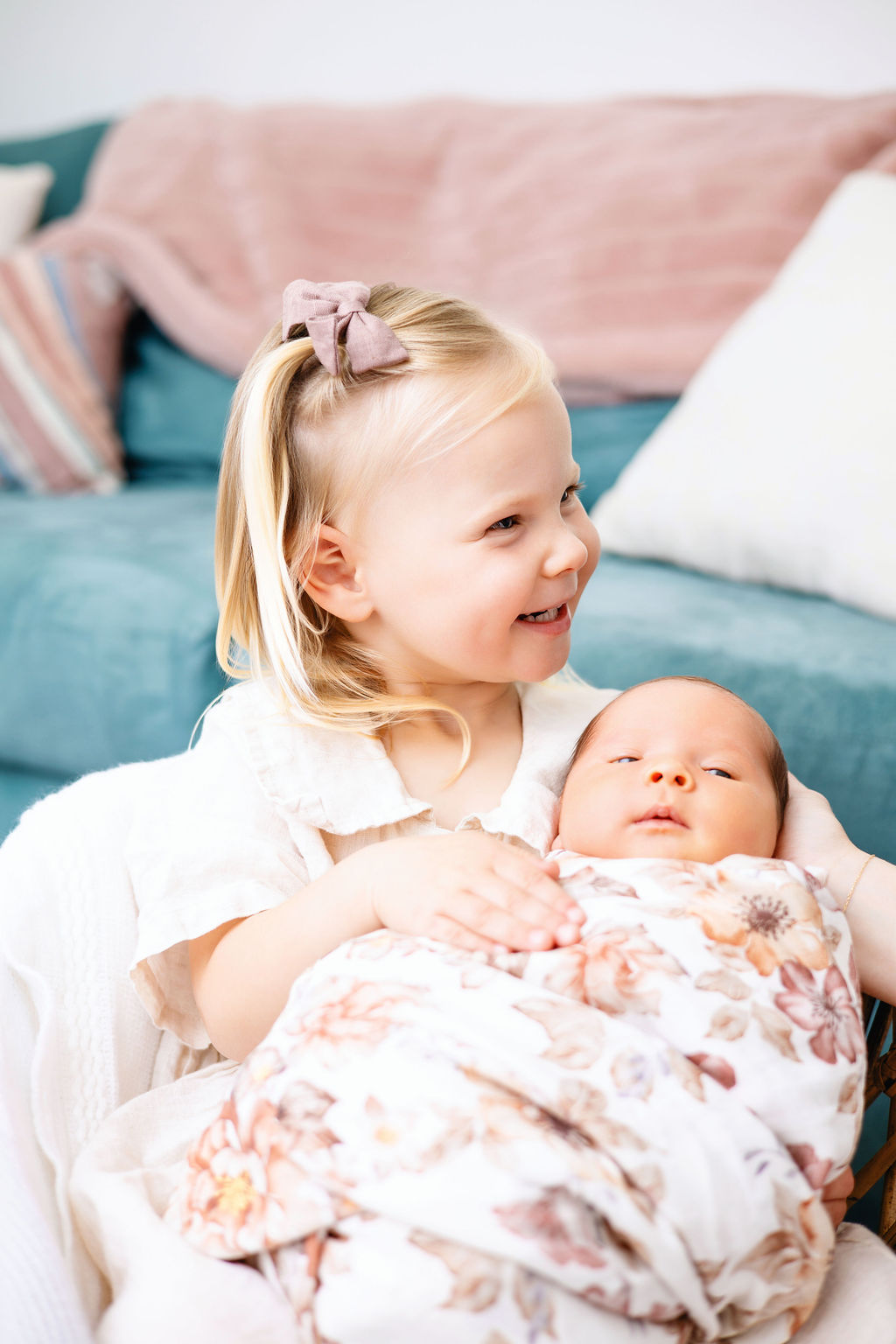 A smiling toddler girl in a white dress sits with her awake newborn baby sister in her lap after meeting postpartum doulas in austin