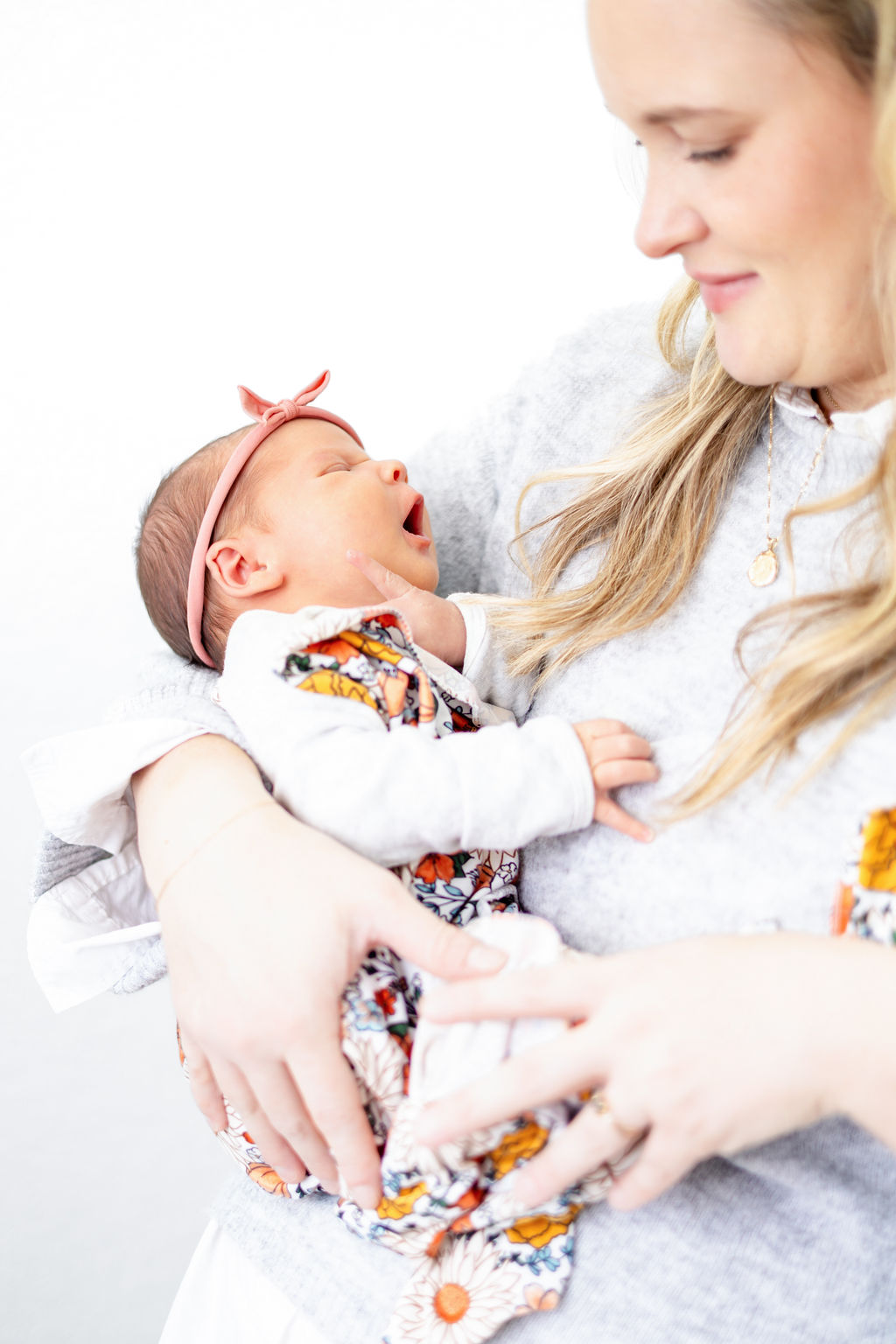 A happy mom in a grey sweater smiles while holding her yawning newborn in a colorful onesie thanks to a postpartum doulas in austin