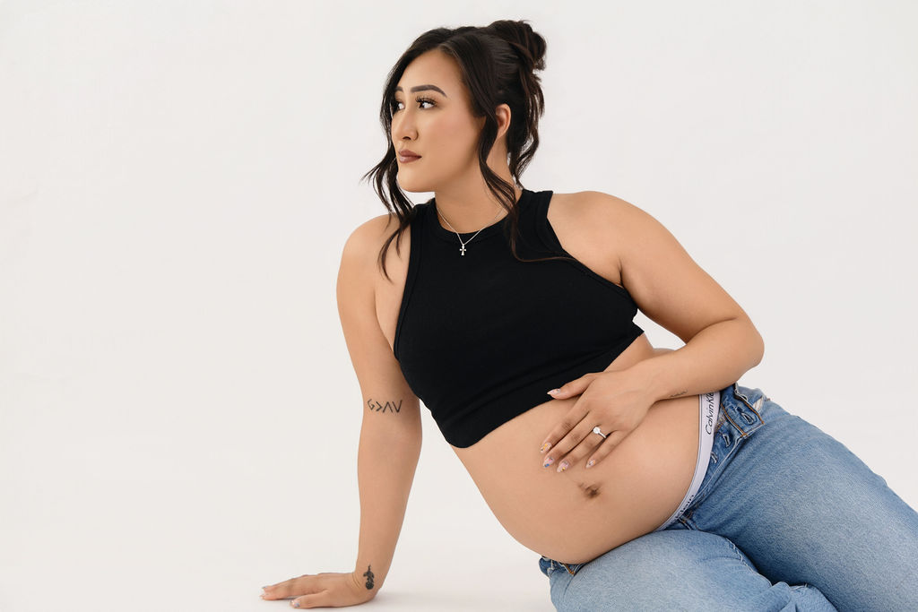 A smiling mom to be sits on the floor of a white studio with a hand on her bump and smiling off to the distance