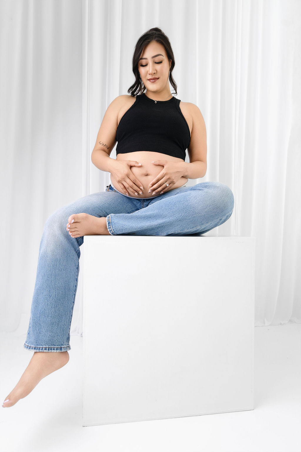A mother to be holds her hands on her bump while sitting on a white cube in a studio after enjoying a prenatal massage in austin