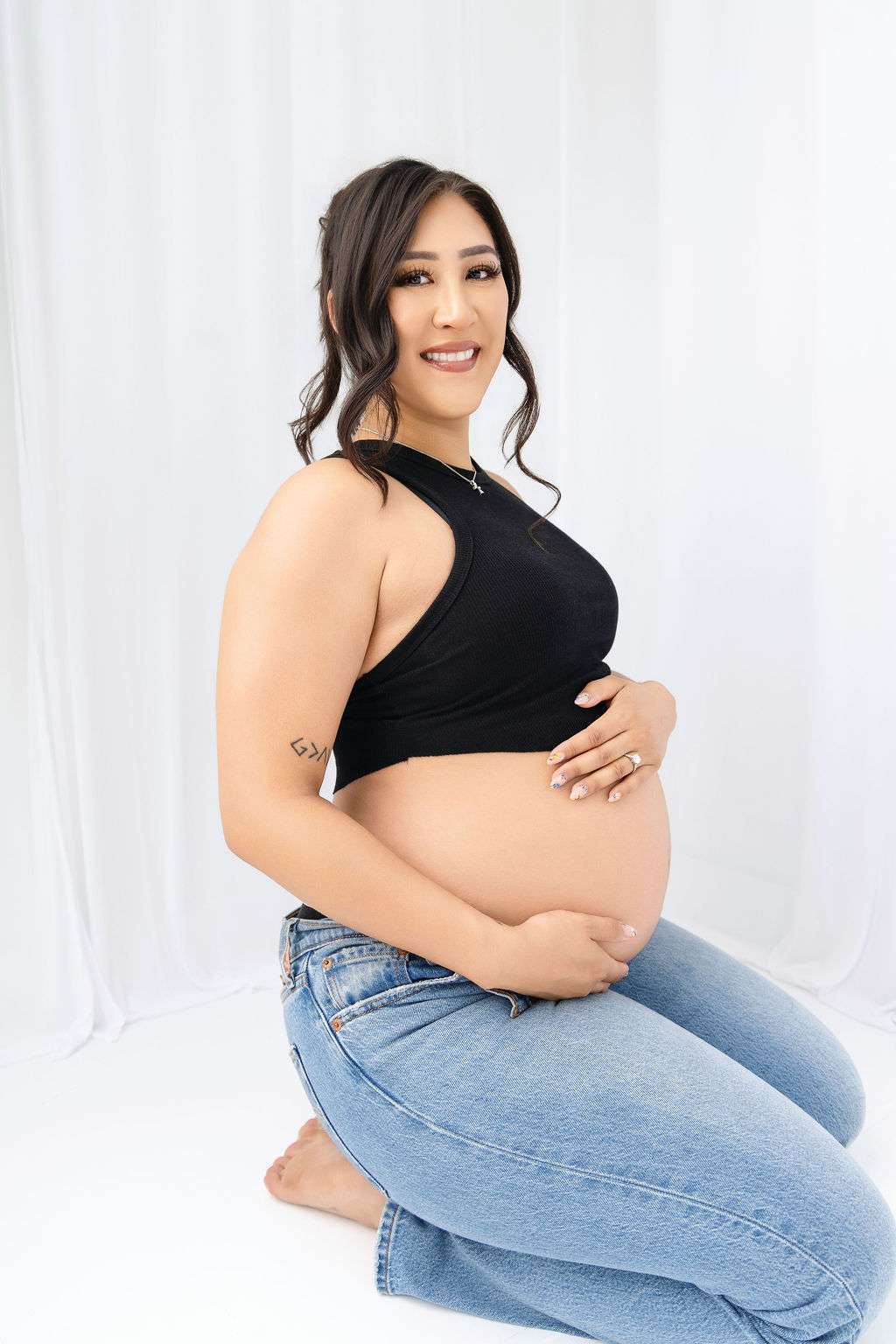 A smiling pregnant woman kneels in a studio holding her bump in a black top and jeans after a prenatal massage in austin