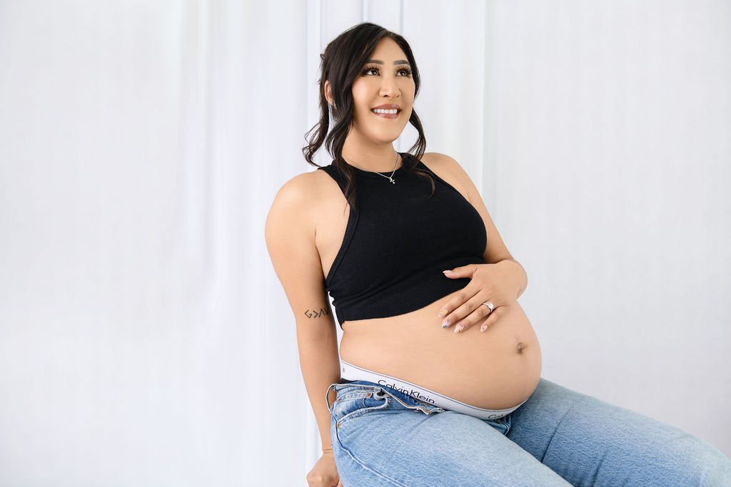 A smiling mother to be sits in a studio in a black blouse and jeans with the bump out