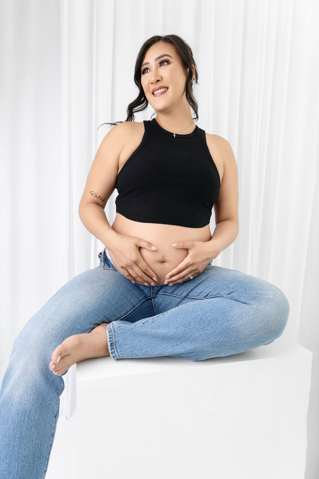 A woman in a black top and jeans smiles while holding her exposed bump in a studio on a white block after some prenatal yoga in austin