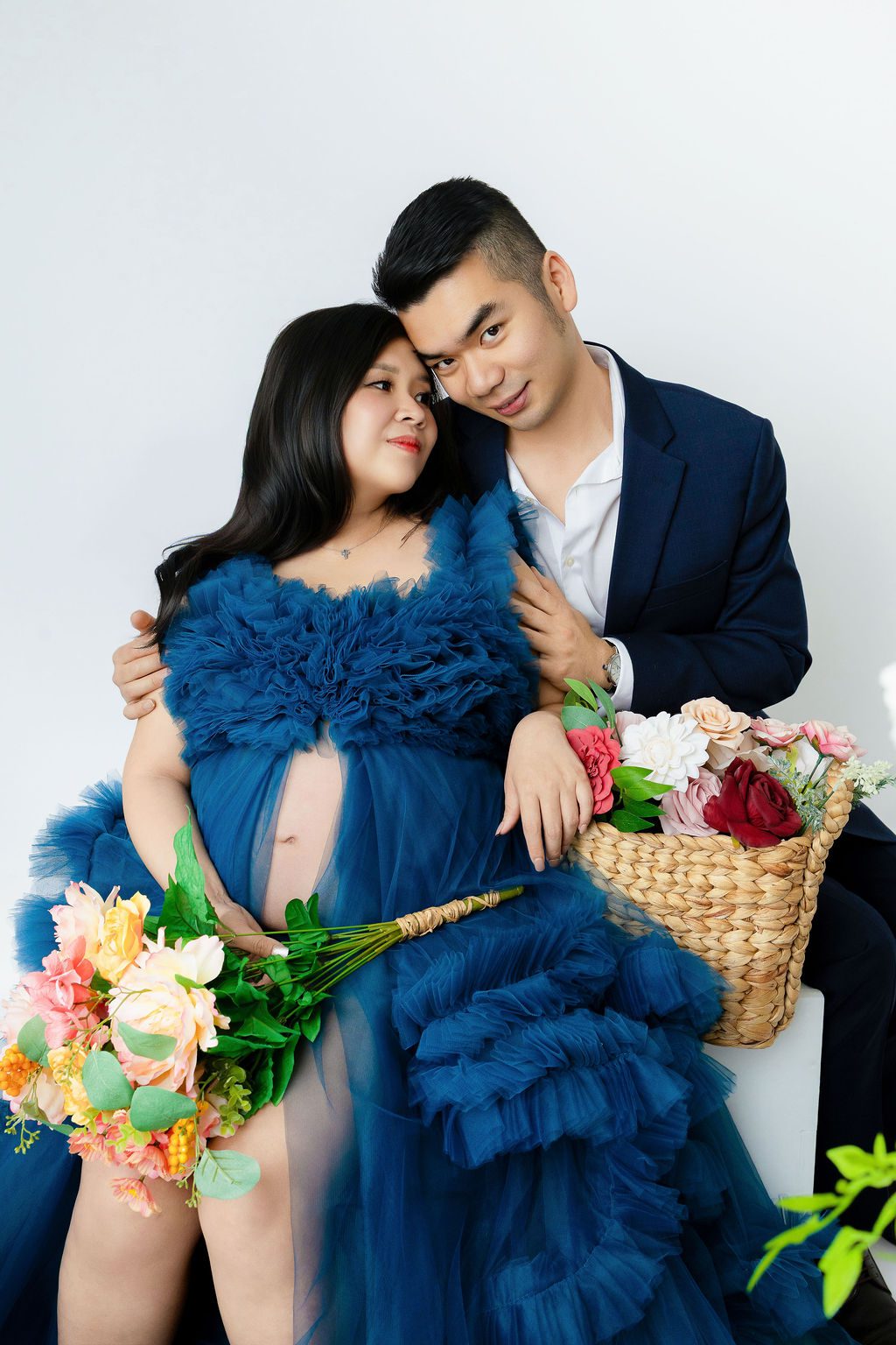 A happy couple snuggles in a studio in blue outfits with pup out holding colorful flowers after visiting baby shower venues in austin