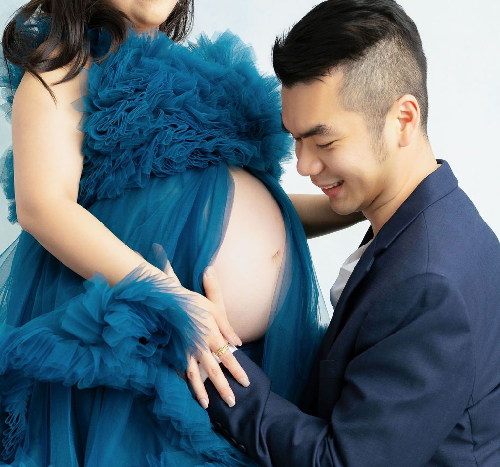 A happy dad to be in a blue suit kneels and smiles t mom's exposed bump in a maternity gown in a studio