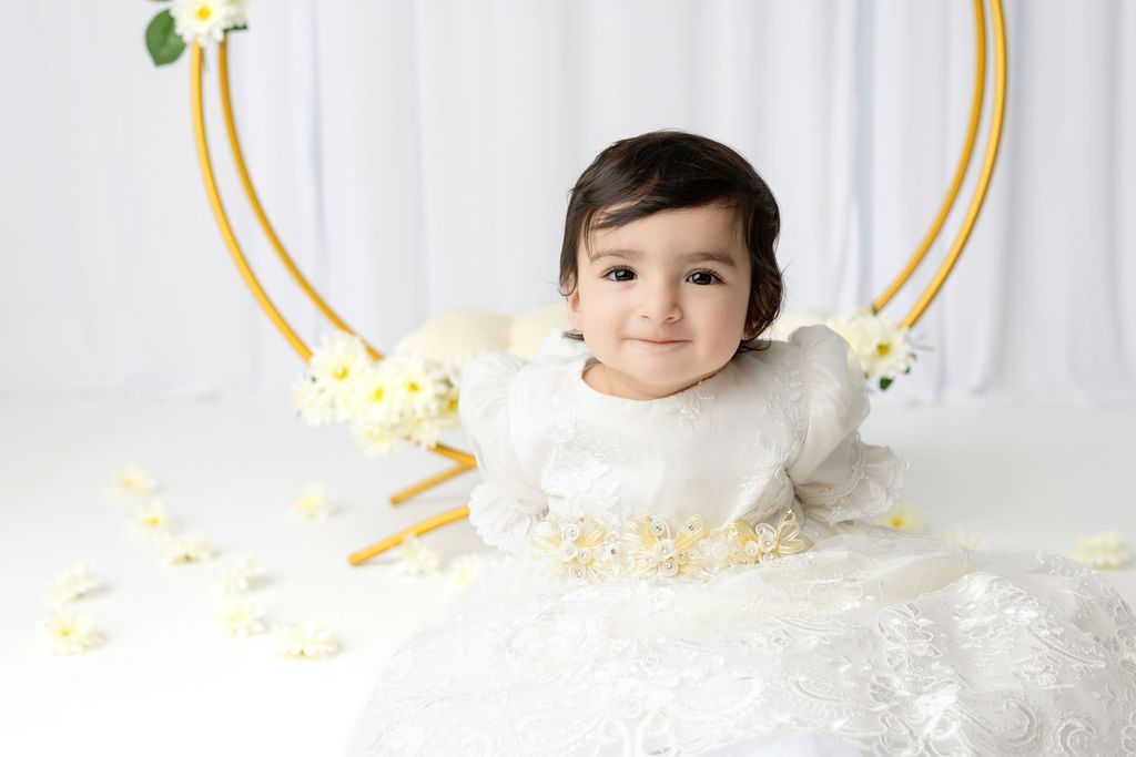 A happy baby girl sits in a studio with white flowers in a white lace gown
