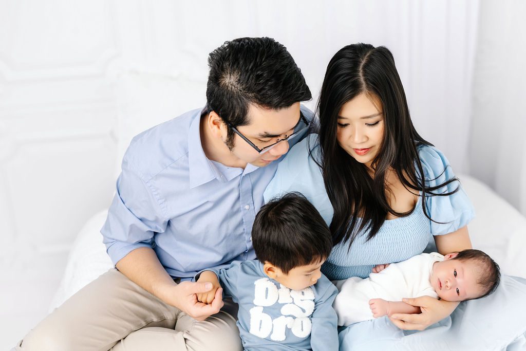 A toddler in mom and dad's laps on a bed looks over at his awake newborn sibling in mom's arms