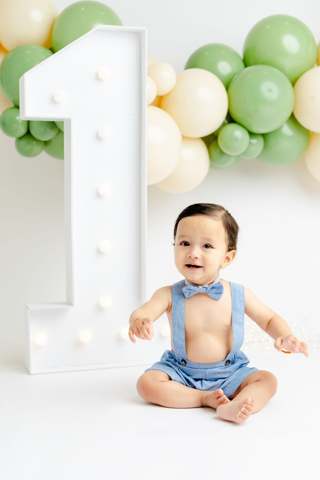 A happy toddler in blue overalls sits with a marquee 1 in a studio under balloons after meeting pediatric dentists in austin