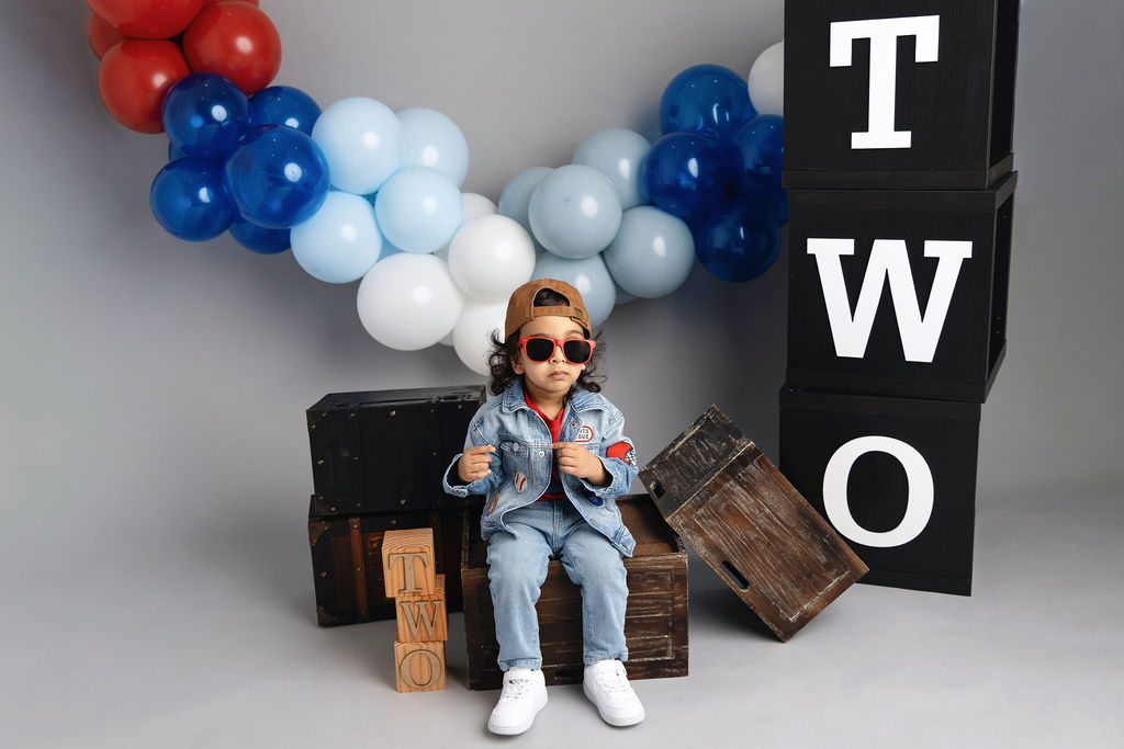 A toddler in denim pants and jacket with a backwards hat and sunglasses sits on apple boxes in a studio