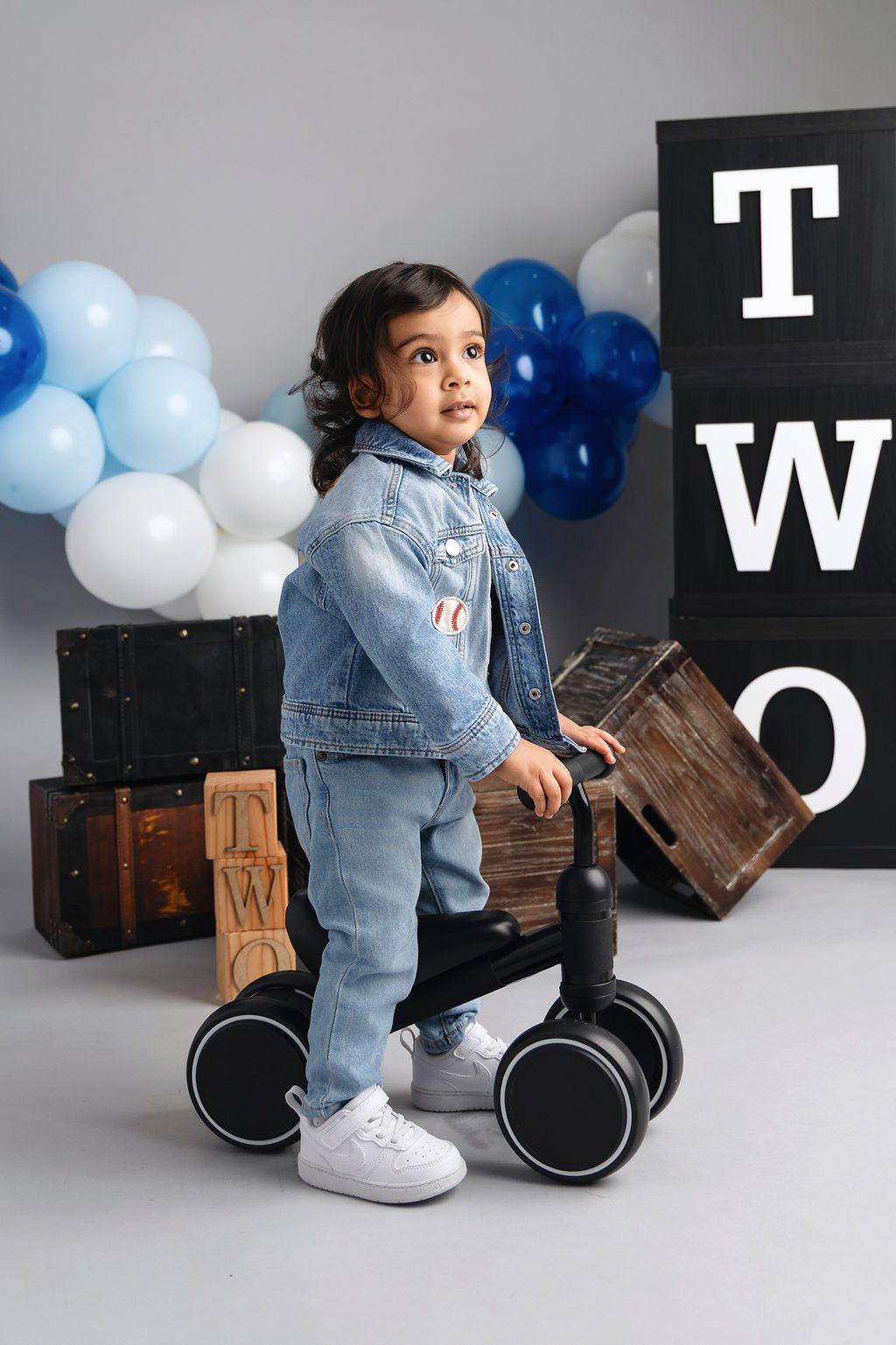 A toddler rides a tricycle in a denim outfit in a Two themed studio after meeting pediatricians in austin