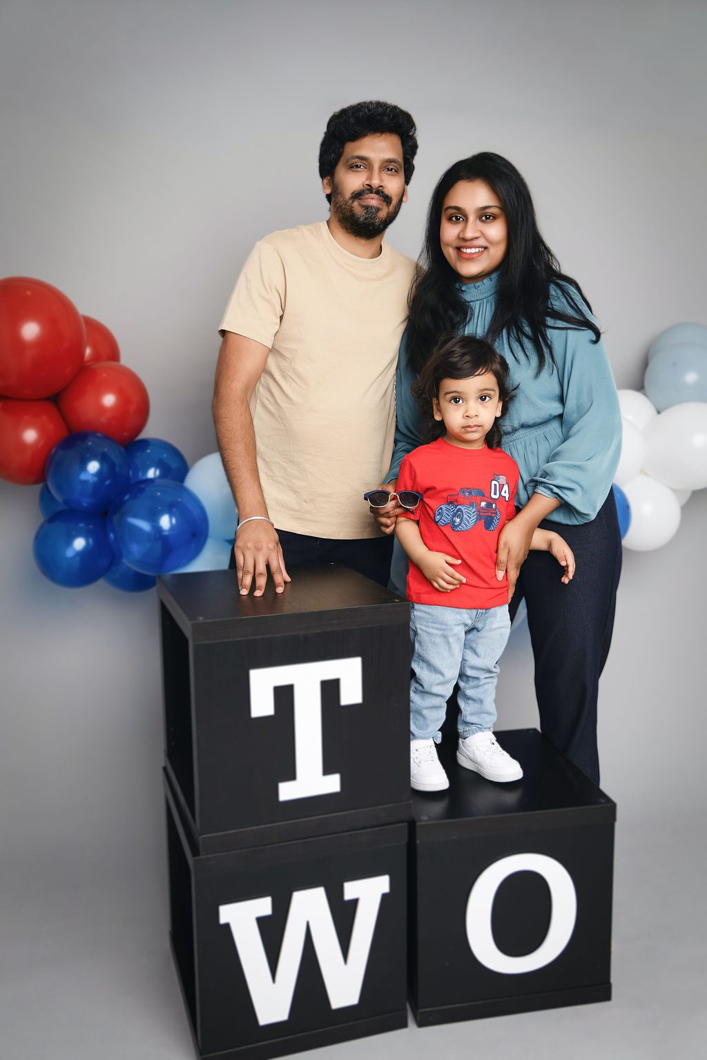 A happy mom and dad stand behind black boxes spelling TWO with their son standing on one in a studio after meeting pediatricians in austin