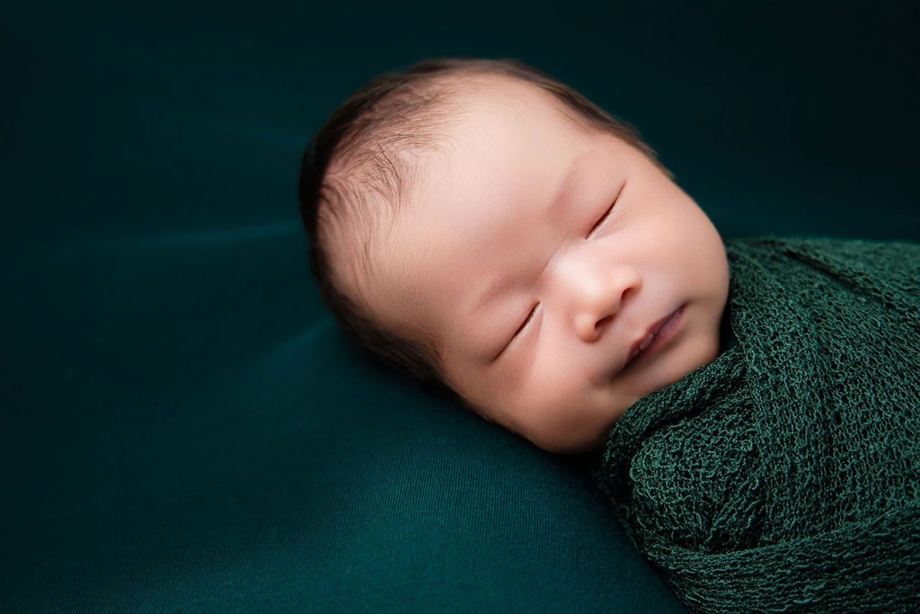 A newborn bay sleeps on his side while wrapped in a knit green swaddle in a studio after using diaper service in austin