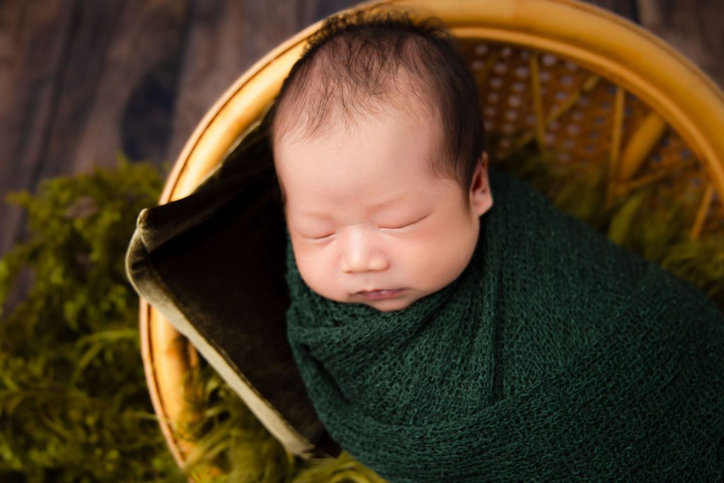 A newborn baby sleeps in a green knit swaddle in a wicker basket thanks to cloth diaper service in austin