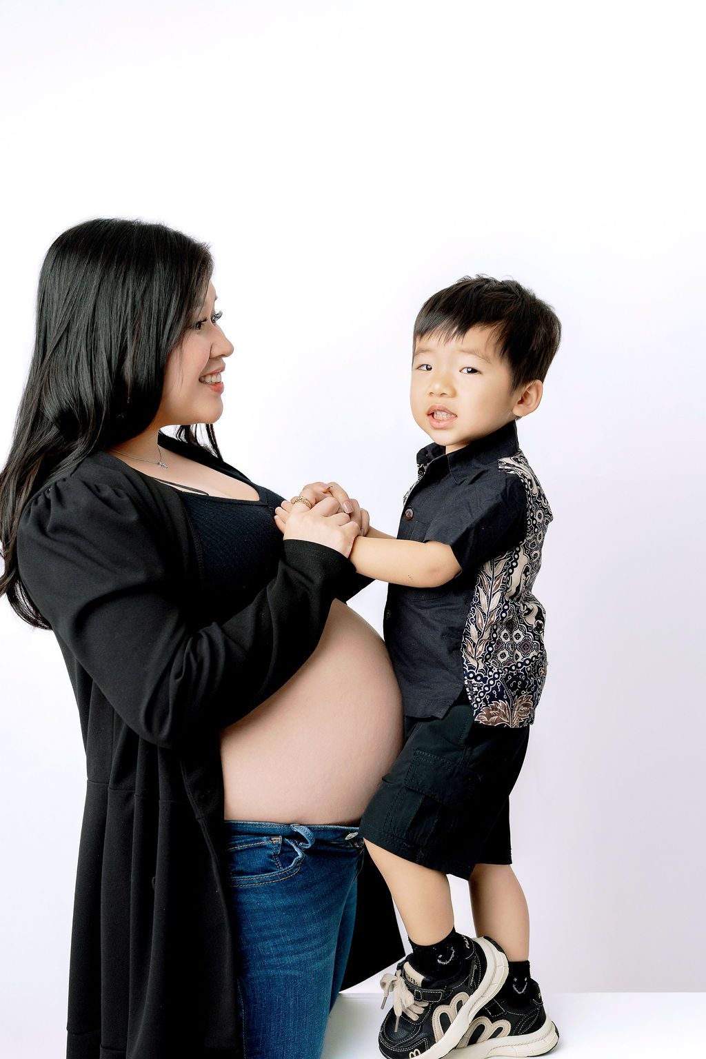 a happy pregnant mother plays holding hands with her toddler son while they both wear black in a studio