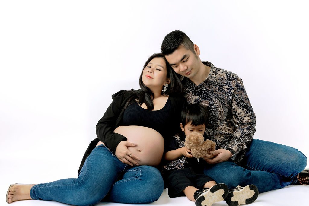 A happy pregnant couple snuggle on the floor while playing with toddler between them in jeans and black tops thanks to fertility clinics in austin